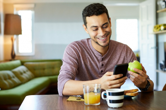 Un jeune homme souriant regarde son téléphone en dînant.