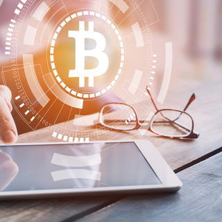 Un logo Bitcoin placé au-dessus de la main d’un homme qui utilise une tablette, à proximité d’une paire de lunettes et d’une tasse de café.